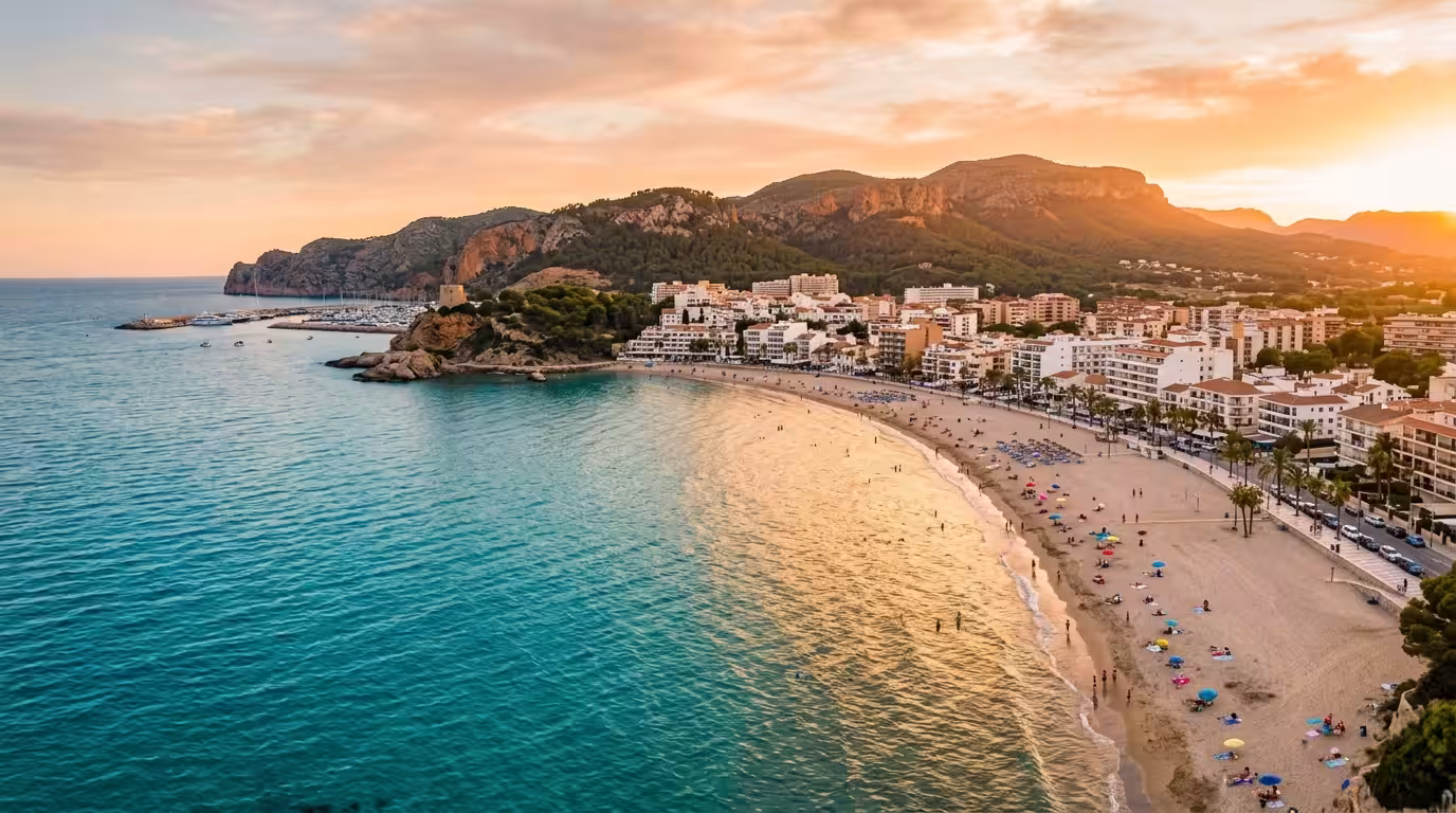 A wide cinematic aerial shot of Oropesa del Mar at golden hour, showing the turquoise Mediterranean Sea meeting a curved sandy beach, with whitewashed buildings and pine-covered cliffs in the backgrou