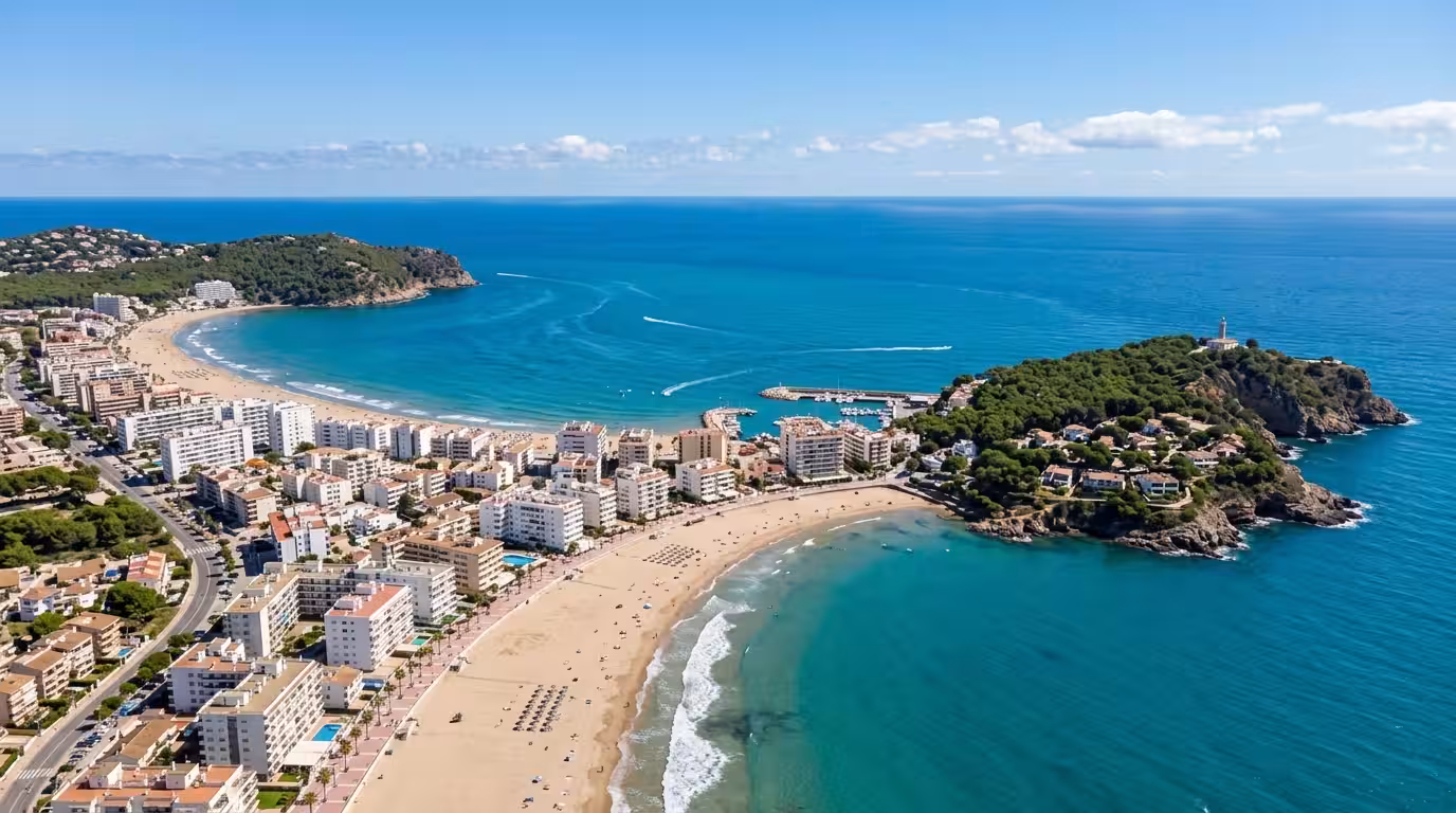 An aerial view of the coastline of Oropesa del Mar on a clear blue-sky afternoon, showing the long sandy beach curving along the Mediterranean, with white apartment buildings and holiday rentals linin