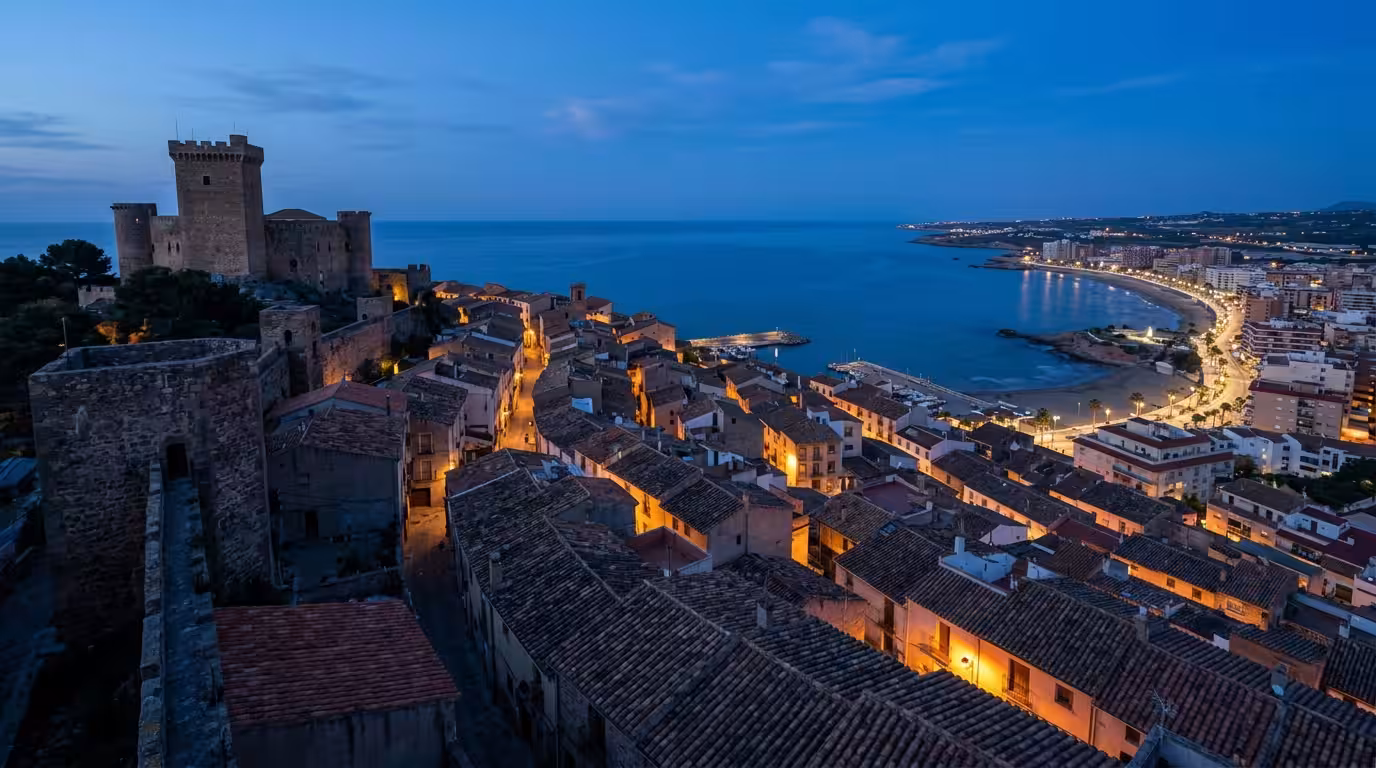 A cool blue-hour photograph taken from the medieval old town of Oropesa del Mar (Orpesa), Spain, looking down over the terracotta rooftops and the ancient stone castle tower toward the Mediterranean S