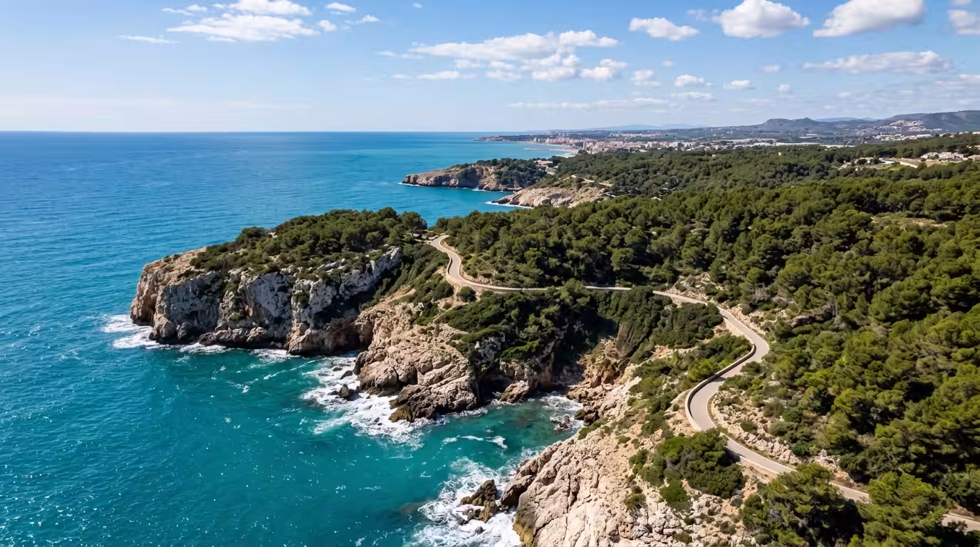 A wide cinematic aerial shot of the Costa de Castellón coastline near Oropesa del Mar, showing rugged limestone cliffs dropping into clear turquoise Mediterranean water, a narrow coastal cycling path 