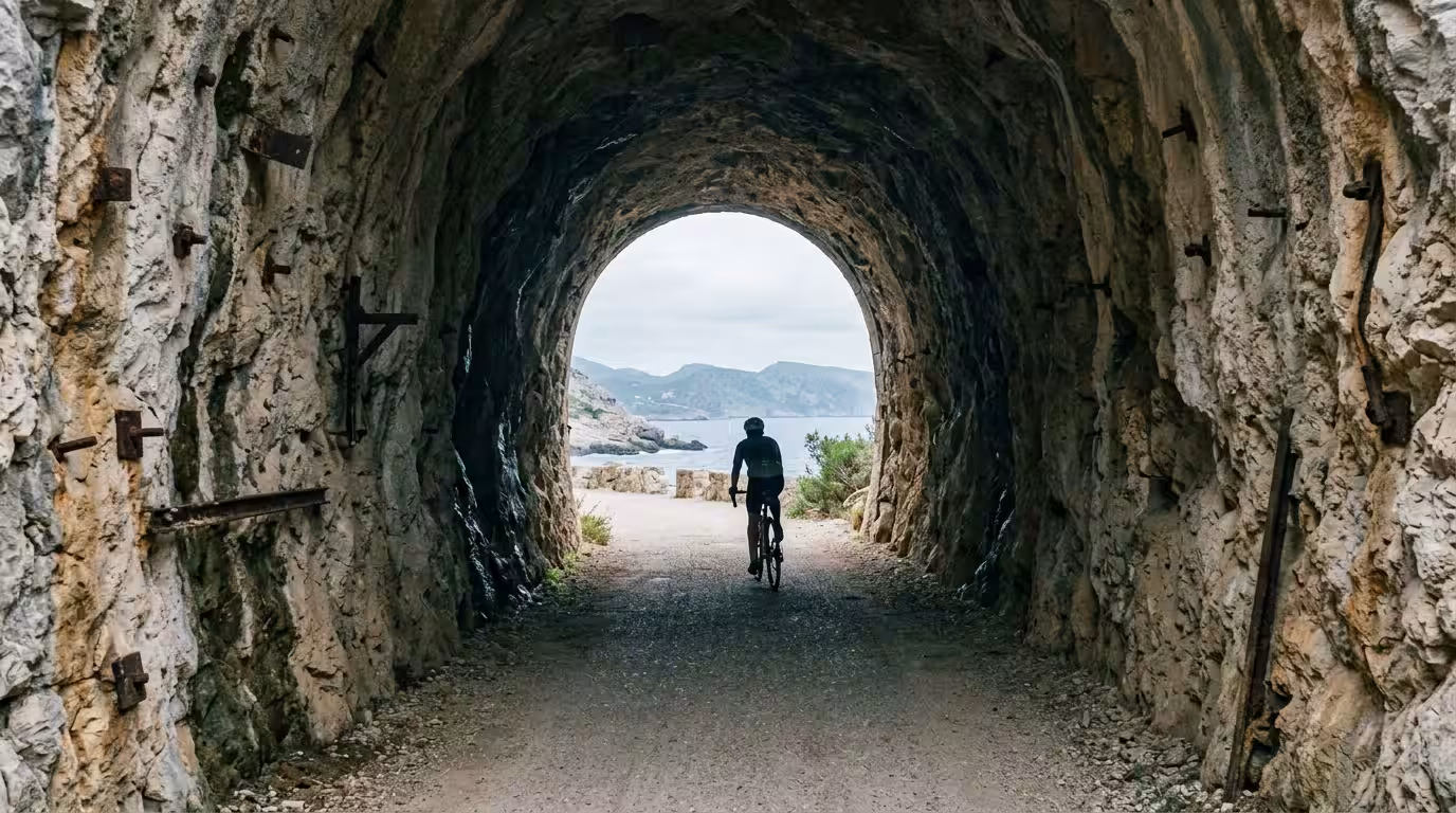 A ground-level documentary shot taken inside a rock-cut tunnel on the Vía Verde del Mar, looking toward the circular exit where bright Mediterranean daylight floods in, the tunnel walls rough-hewn lim