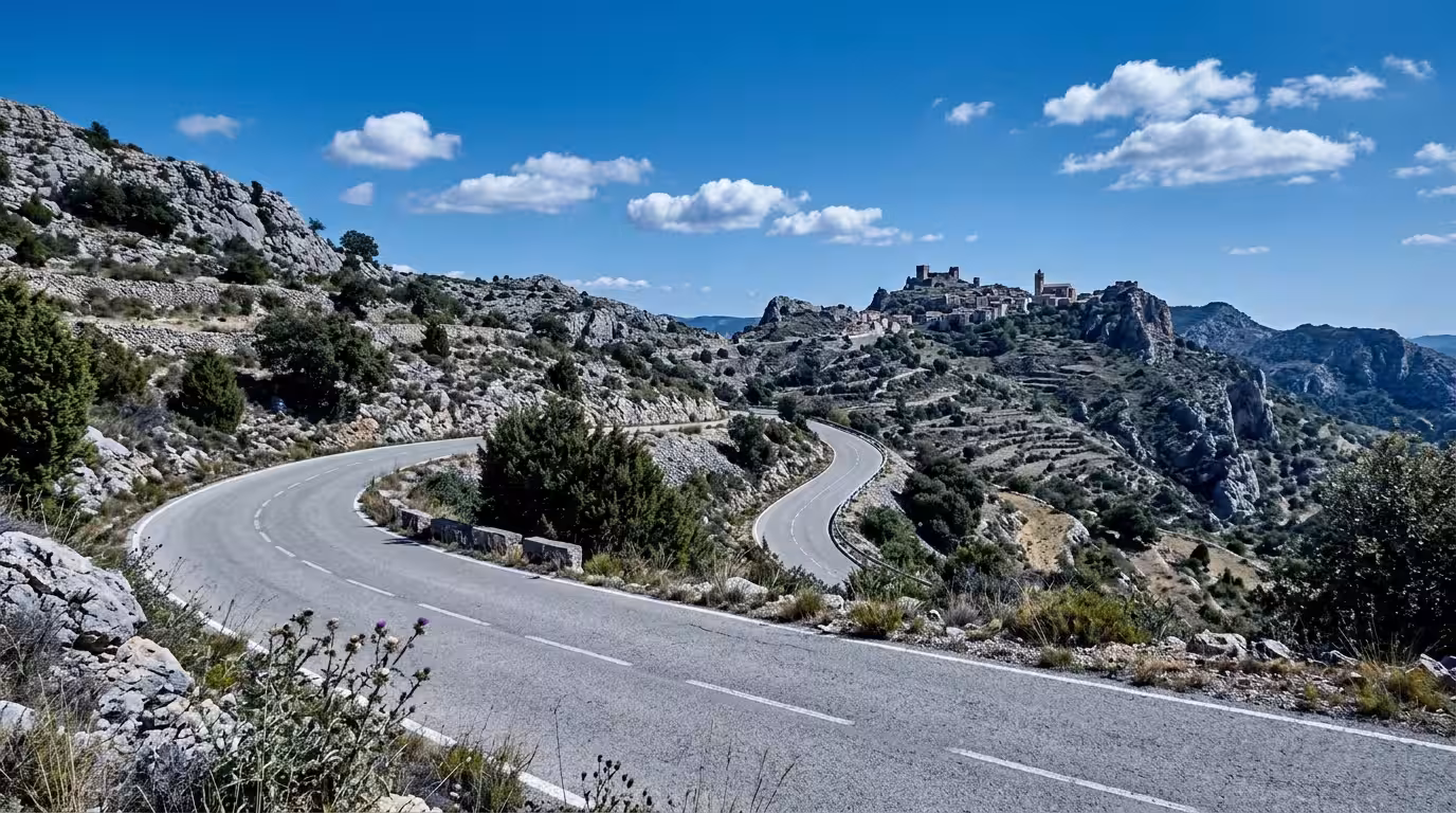 A narrow, winding mountain road cutting through the rocky limestone landscape of the Alt Maestrat in Castellón, Spain. It is midday with bright, clear Mediterranean light. Sparse juniper and holm oak 