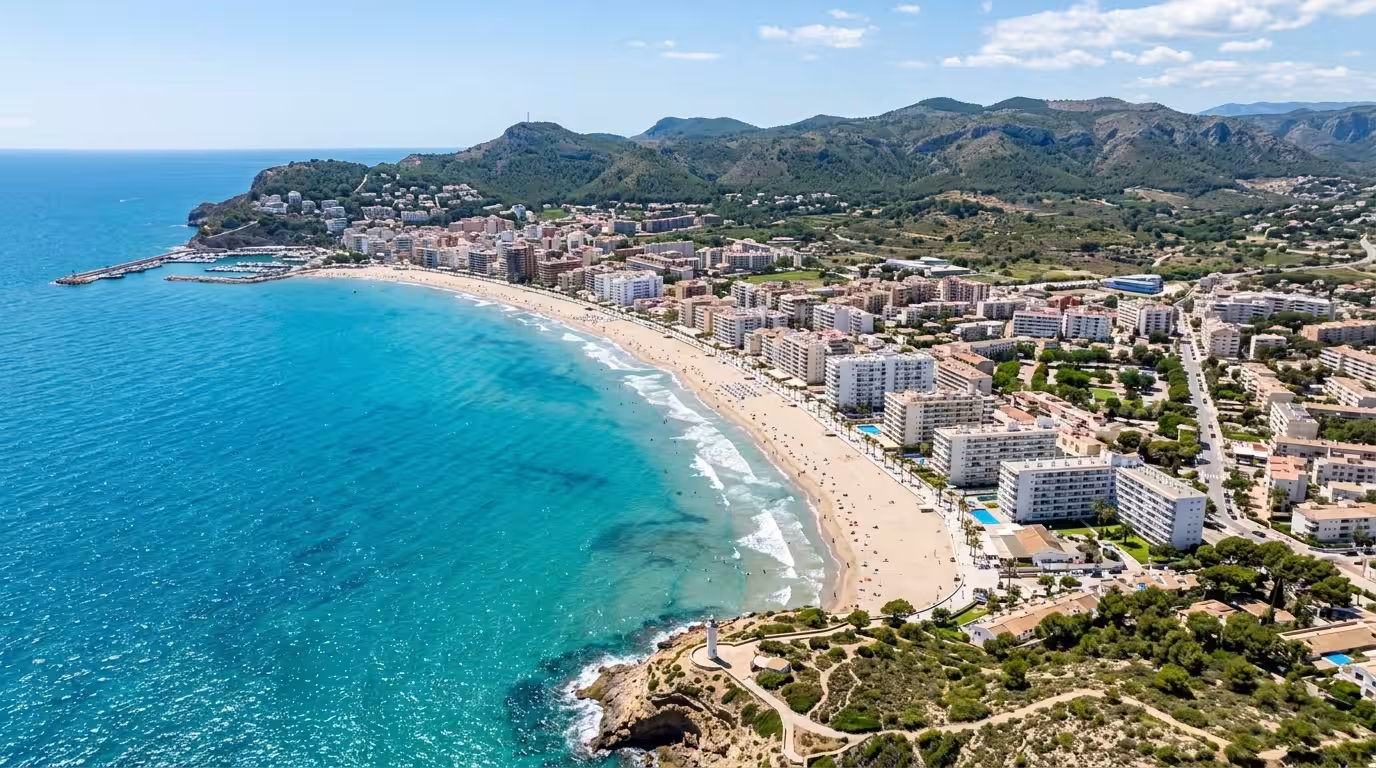 A bright midday aerial view of the coastline of Oropesa del Mar showing the curved sandy beach, turquoise shallow water, the white apartment buildings of the seafront, and the green hilly interior str