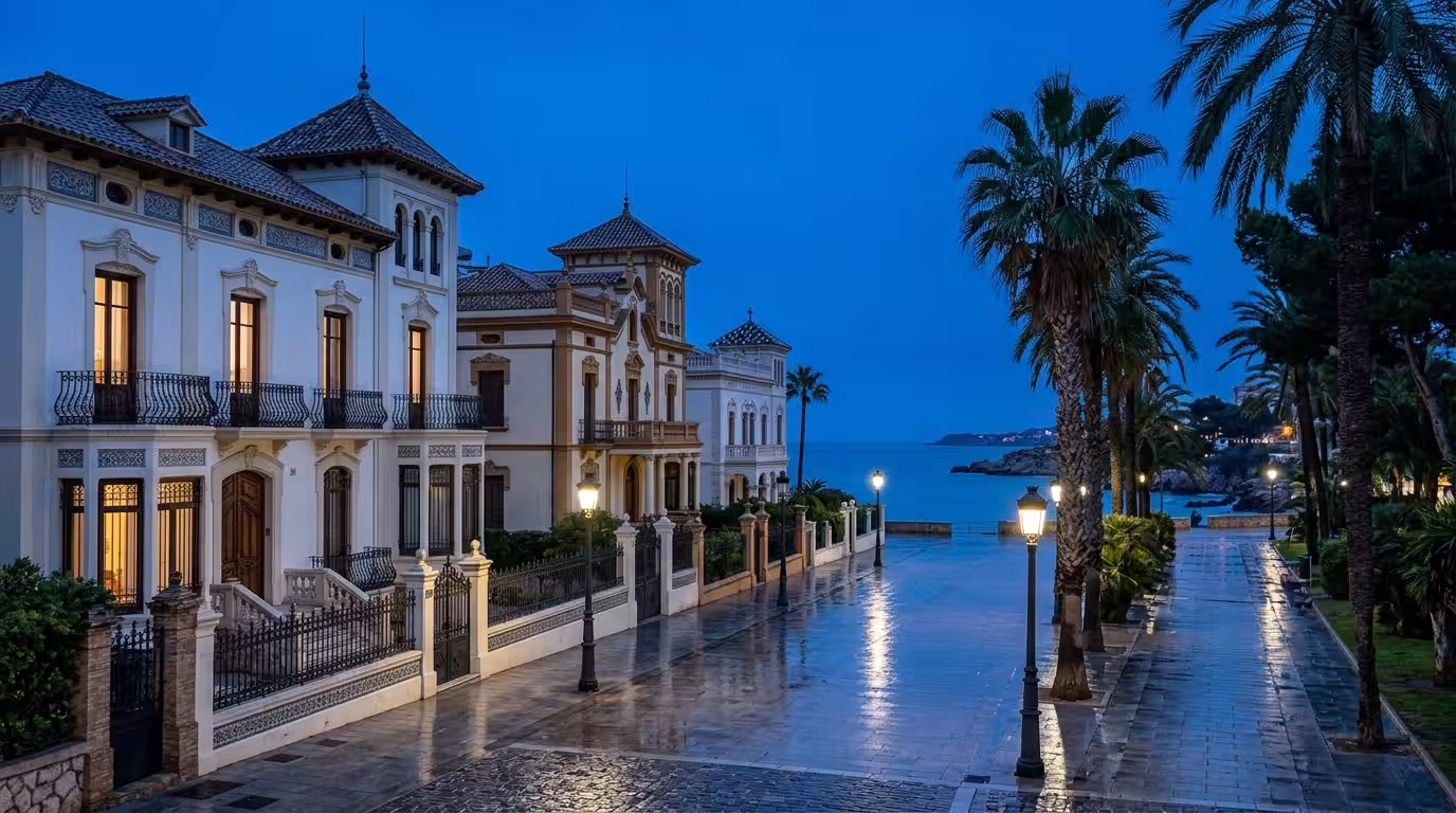A crisp blue-hour photograph of the Benicàssim modernist villas promenade, showing the ornate facades of historic summer mansions in white and ochre with wrought-iron balconies, reflected in the wet p