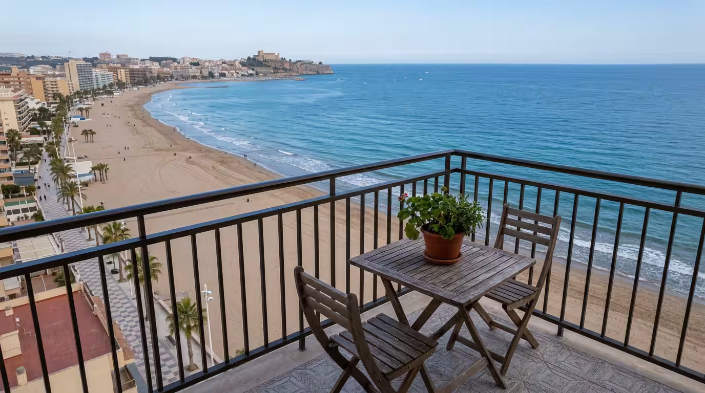 Terraza de un apartamento vacacional en primera línea de playa de Oropesa del Mar al final de la tarde, con una mesa de madera, dos sillas, una planta en maceta y vista directa al mar y a la arena; lu