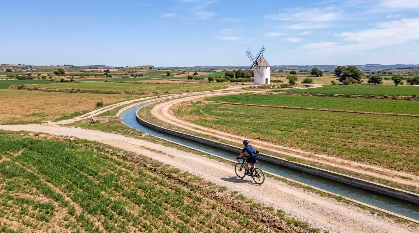 Un ciclista con una cámara colgada al hombro pedalea por un sendero junto a una acequia entre campos de cultivo en el interior de Cabanes, con un molino blanqueado de fondo y el cielo azul claro cubri