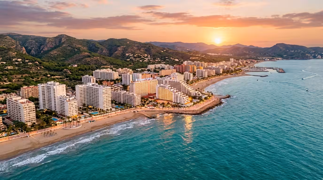 A sweeping golden-hour aerial view of the Oropesa del Mar coastline, showing the white high-rise apartment buildings of the Marina d'Or area reflecting warm sunset light, with turquoise Mediterranean 