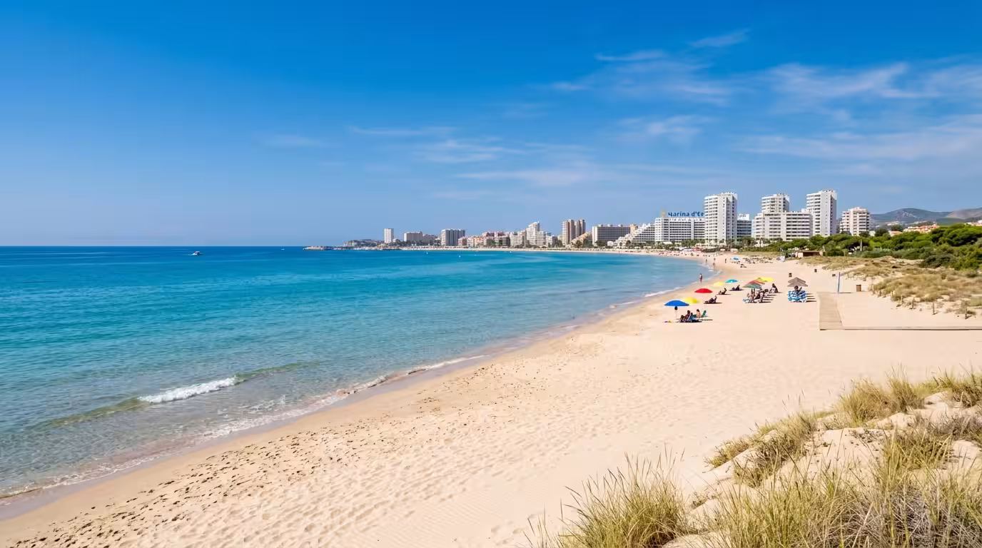 A quiet, sun-drenched Mediterranean beach in early September with calm turquoise water, fine white sand, a few colorful beach umbrellas in the distance, and the characteristic white apartment towers o
